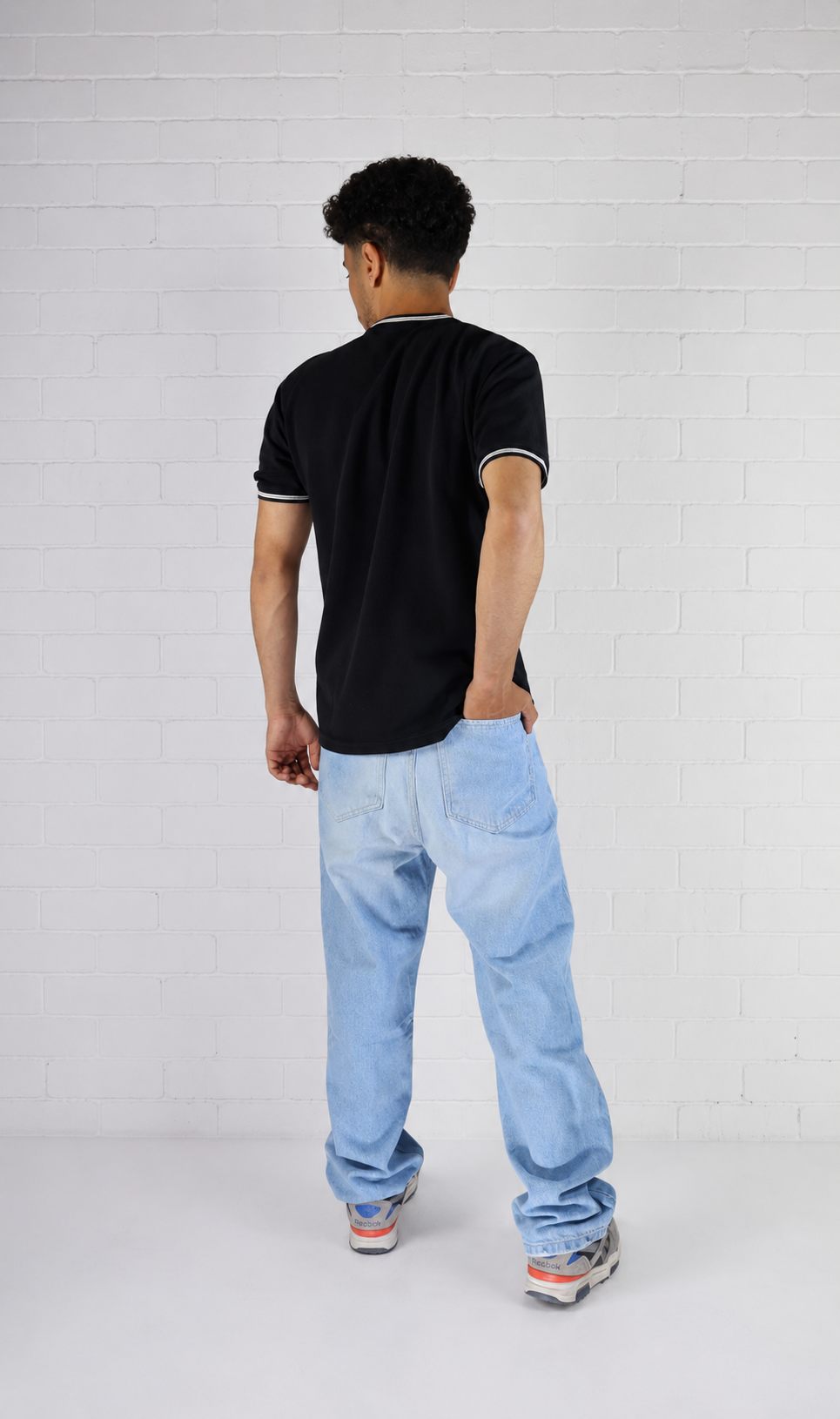 Person wearing a black t-shirt and light blue jeans standing against a white brick wall.